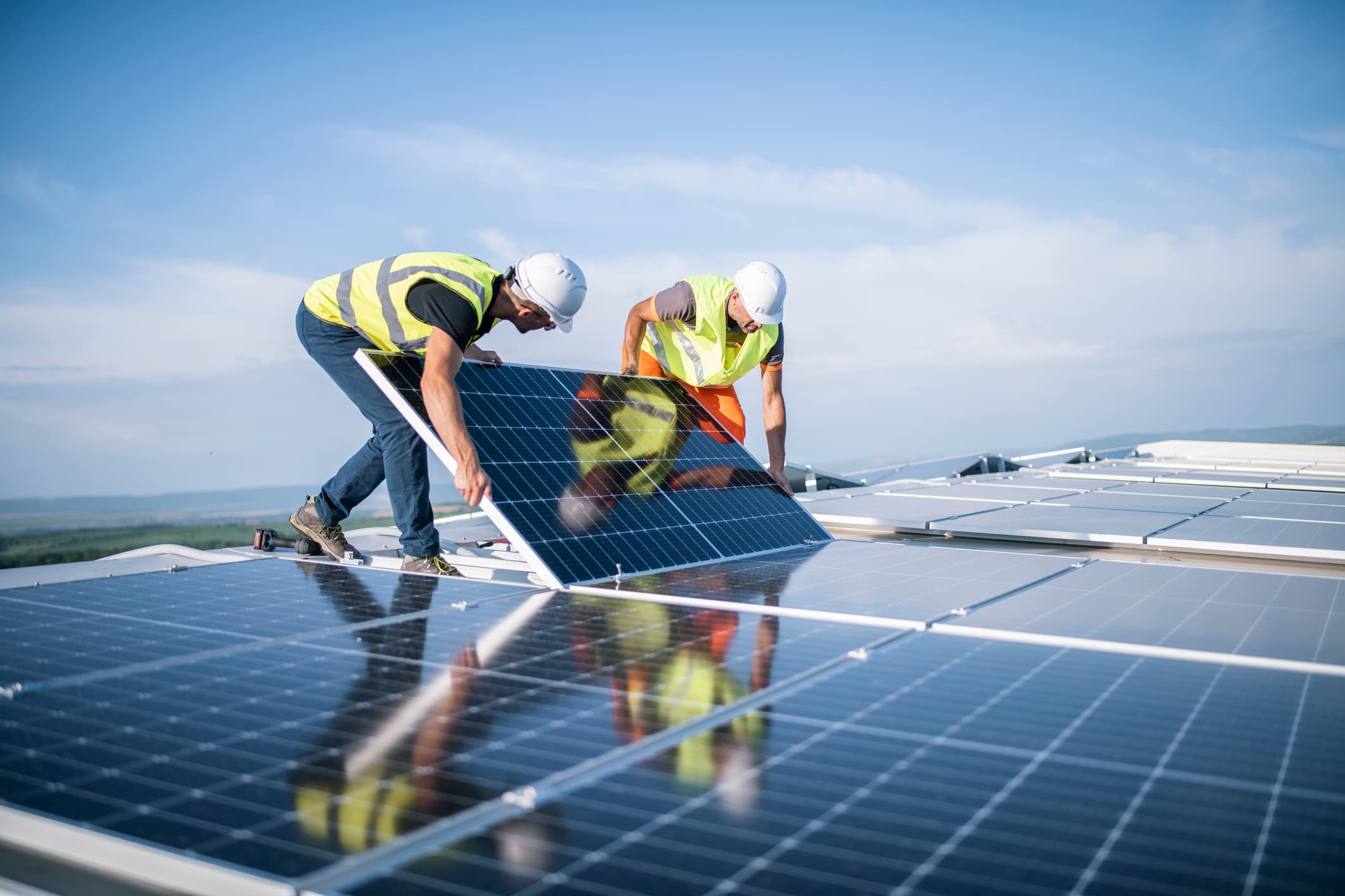 Two workers in hard hats and safety vests installing a solar panel on a rooftop array under a clear blue sky